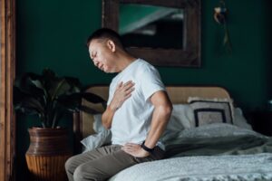 Senior Asian man with eyes closed holding his chest in discomfort, suffering from chest pain while sitting on bed at home. Elderly and health issues concept