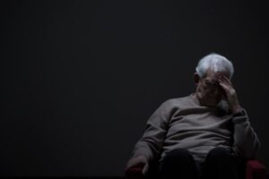A black and white photo of a man sitting with his head in his hands, looking down. His posture and body language suggest he is experiencing a moment of deep sadness or distress.