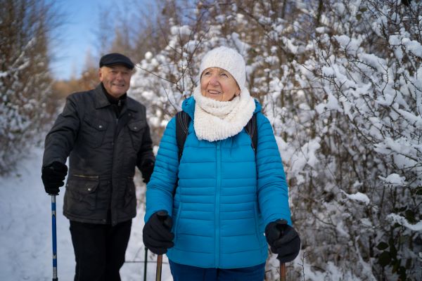 Two older adults are all bundled up in winter gear taking a hike through a snowy forest